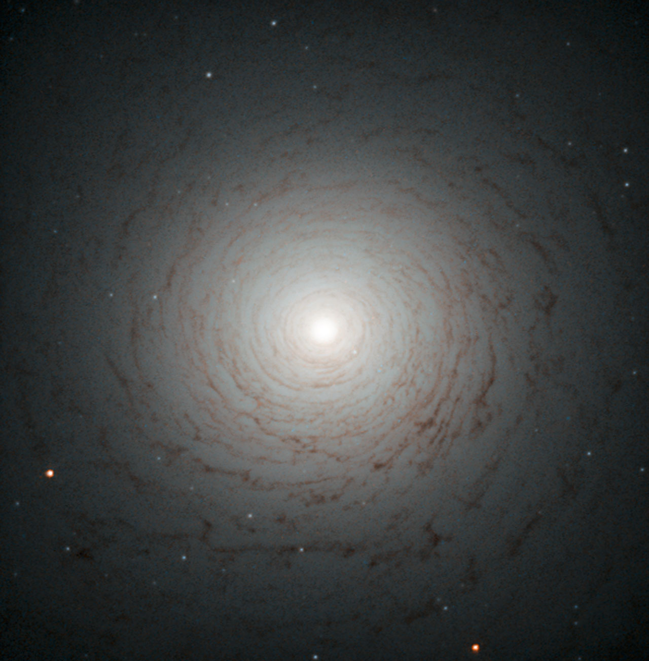 This striking cosmic whirl is the centre of galaxy NGC 524, as seen with the NASA/ESA Hubble Space Telescope. This galaxy is located in the constellation of Pisces, some 90 million light-years from Earth. NGC 524 is a lenticular galaxy. Lenticular galaxies are believed to be an intermediate state in galactic evolution — they are neither elliptical nor spiral. Spirals are middle-aged galaxies with vast, pinwheeling arms that contain millions of stars. Along with these stars are large clouds of gas and dust that, when dense enough, are the nurseries where new stars are born. When all the gas is either depleted or lost into space, the arms gradually fade away and the spiral shape begins to weaken. At the end of this process, what remains is a lenticular galaxy — a bright disc full of old, red stars surrounded by what little gas and dust the galaxy has managed to cling on to. This image shows the shape of NGC 524 in detail, formed by the remaining gas surrounding the galaxy’s central bulge. Observations of this galaxy have revealed that it maintains some spiral-like motion, explaining its intricate structure. A version of this image was entered into the Hubble’s Hidden Treasures image processing competition by contestant Judy Schmidt.
