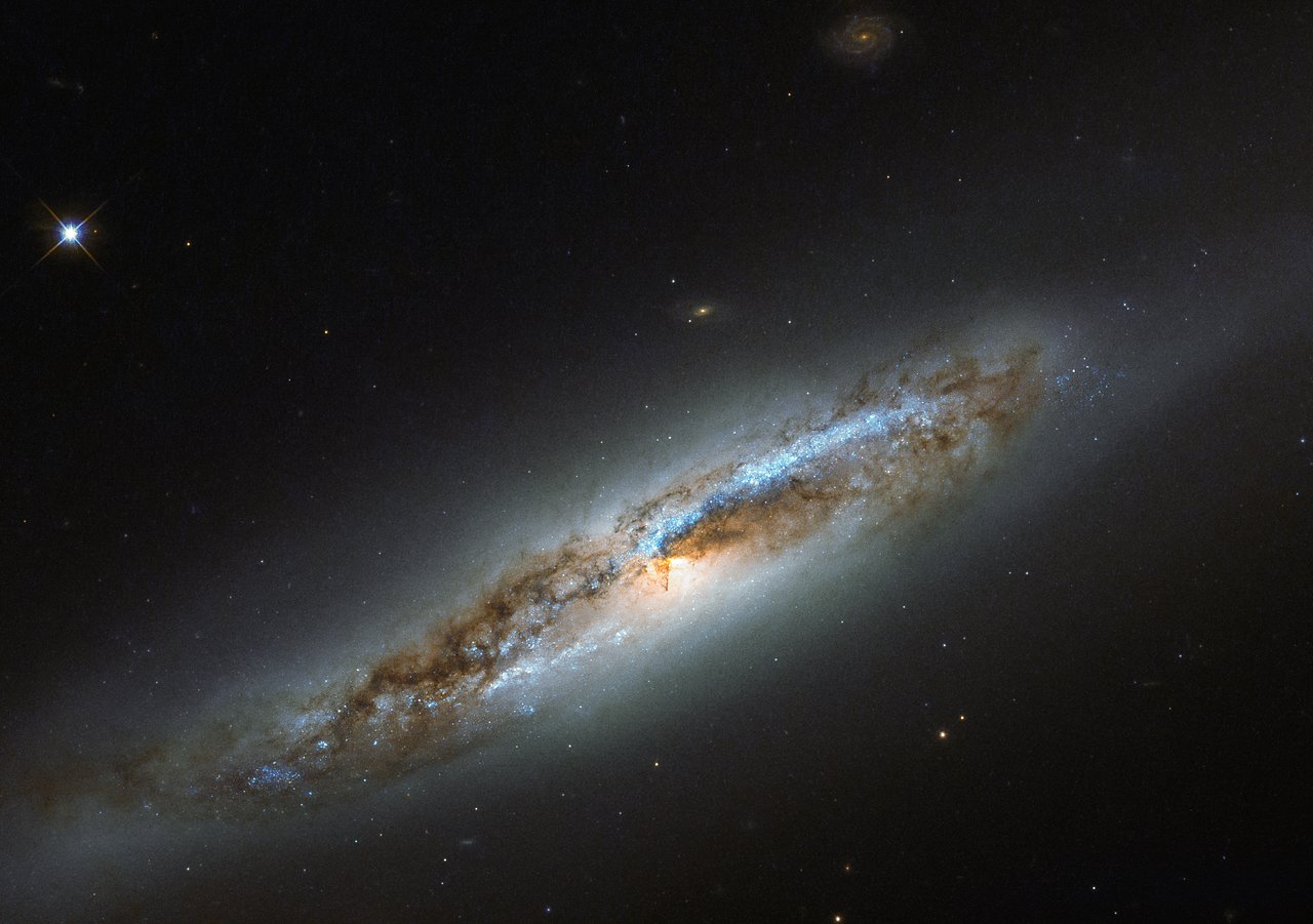 The constellation of Virgo (The Virgin) is especially rich in galaxies, due in part to the presence of a massive and gravitationally-bound collection of over 1300 galaxies called the Virgo Cluster. One particular member of this cosmic community, NGC 4388, is captured in this image, as seen by the NASA/ESA Hubble Space Telescope’s Wide Field Camera 3 (WFC3). Located some 60 million light-years away, NGC 4388 is experiencing some of the less desirable effects that come with belonging to such a massive galaxy cluster. It is undergoing a transformation, and has taken on a somewhat confused identity. While the galaxy’s outskirts appear smooth and featureless, a classic feature of an elliptical galaxy, its centre displays remarkable dust lanes constrained within two symmetric spiral arms, which emerge from the galaxy’s glowing core — one of the obvious features of a spiral galaxy. Within the arms, speckles of bright blue mark the locations of young stars, indicating that NGC 4388 has hosted recent bursts of star formation. Despite the mixed messages, NGC 4388 is classified as a spiral galaxy. Its unusual combination of features are thought to have been caused by interactions between NGC 4388 and the Virgo Cluster. Gravitational interactions — from glancing blows to head-on collisions, tidal influencing, mergers, and galactic cannibalism — can be devastating to galaxies. While some may be lucky enough to simply suffer a distorted spiral arm or newly-triggered wave of star formation, others see their structure and contents completely and irrevocably altered.